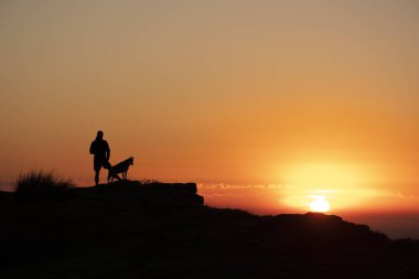 Hiker with dog watching the sunrise from the top of the mountain.