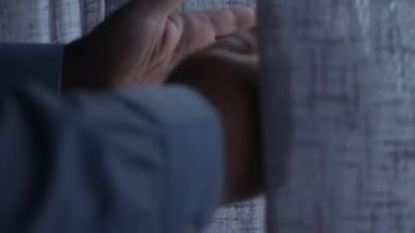 Perdenin Arkasından casusluk yaparak perdeleri kaldırır ve pencereye bakar. Pencere Perdesinden Dışarıya Bakan Bir Kişi.