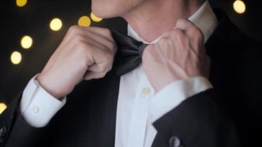 Close up view of man in a suit adjusts his bow tie. Black background with lights.