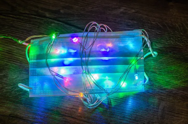 Noel çelenginde koruyucu bir tıbbi maske renkli ışıklarla parlar ve parlar.