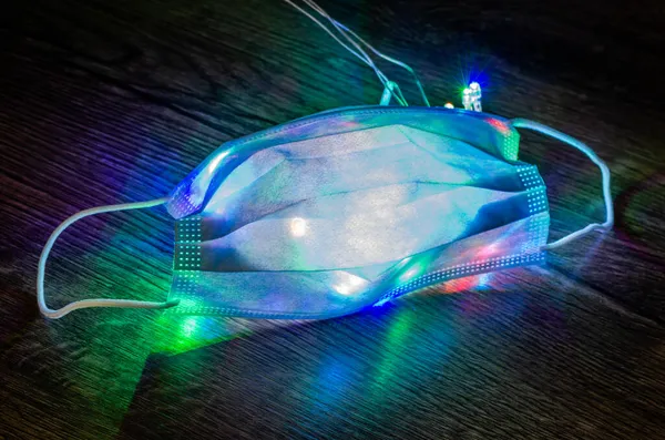 Noel çelenginde koruyucu bir tıbbi maske renkli ışıklarla parlar ve parlar.