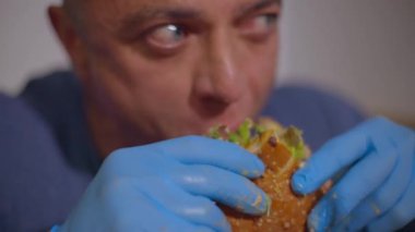İştahlı yetişkin kel bir adam lezzetli bir burgeri eldivenle çiğner, yemeğe odaklanır, kapatır.
