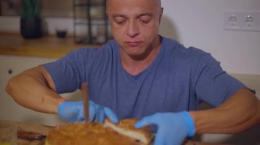 Yetişkin kel bir adam hamburgeri dilimledi ve eldivenli ellerle parçaları yedi.