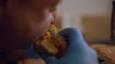 Yetişkin kel bir adam eldivenli sulu bir hamburger yiyor. Her lokmanın tadını çıkarıyor. Yaklaş.