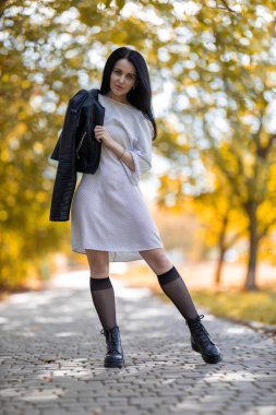 Beyaz elbiseli, deri ceketli, altın sonbahar arkaplanına poz veren güzel esmer kadın. Moda fotoğrafı..