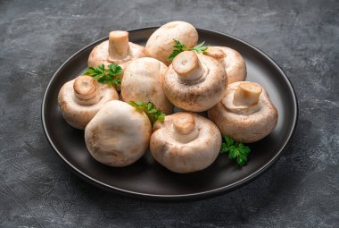 Siyah bir arka planda siyah bir tabakta taze mantar ve maydanoz. Yan görünüm, yakın plan.