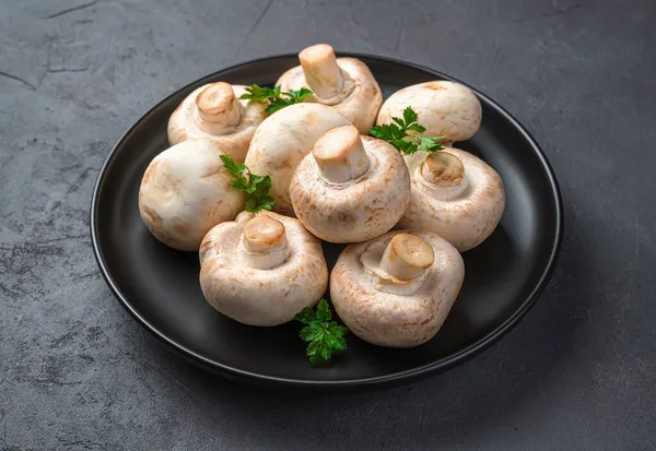 Siyah bir arka planda siyah bir tabakta taze mantar ve maydanoz. Yan görünüm, yakın plan.