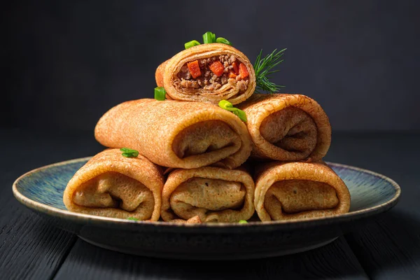 Fried pancakes stuffed with meat and carrots are decorated with fresh onions and dill on a dark background close-up. Side view.
