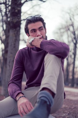 Güz mevsiminde koyu renk saçlı güzel bir İtalyan çocuğu koyu kırmızı kazağıyla bankta oturuyor. Yüksek kalite fotoğraf