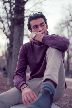 Güz mevsiminde koyu renk saçlı güzel bir İtalyan çocuğu koyu kırmızı kazağıyla bankta oturuyor. Yüksek kalite fotoğraf