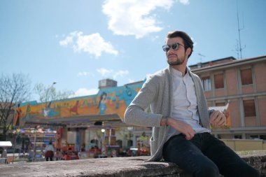 Yakışıklı, siyah saçlı, ceketli, güneşli bir günde alçak duvarda oturan adam. Yüksek kalite fotoğraf