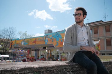 Yakışıklı, siyah saçlı, ceketli, güneşli bir günde alçak duvarda oturan adam. Yüksek kalite fotoğraf