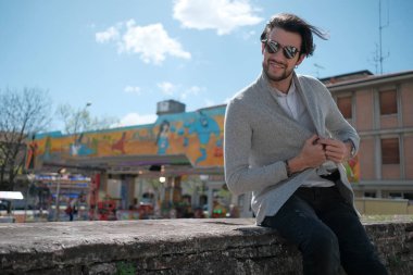 Yakışıklı, siyah saçlı, ceketli, güneşli bir günde alçak duvarda oturan adam. Yüksek kalite fotoğraf