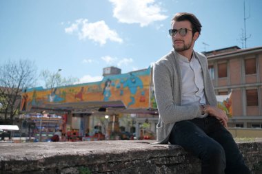 Yakışıklı, siyah saçlı, ceketli, güneşli bir günde alçak duvarda oturan adam. Yüksek kalite fotoğraf