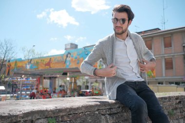 Yakışıklı, siyah saçlı, ceketli, güneşli bir günde alçak duvarda oturan adam. Yüksek kalite fotoğraf