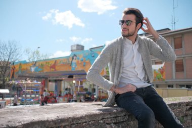 Yakışıklı, siyah saçlı, ceketli, güneşli bir günde alçak duvarda oturan adam. Yüksek kalite fotoğraf