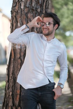 Yakışıklı, siyah saçlı, güneş gözlüklü, geniş çam gövdesine yaslanmış bir çocuk. Yüksek kalite fotoğraf