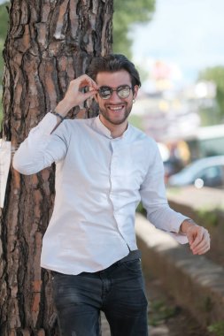 Yakışıklı, siyah saçlı, güneş gözlüklü, geniş çam gövdesine yaslanmış bir çocuk. Yüksek kalite fotoğraf