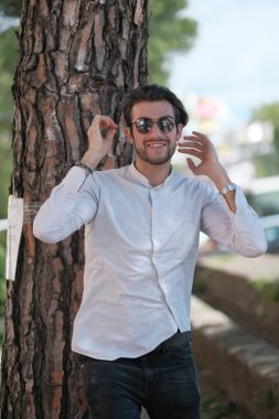 Yakışıklı, siyah saçlı, güneş gözlüklü, geniş çam gövdesine yaslanmış bir çocuk. Yüksek kalite fotoğraf