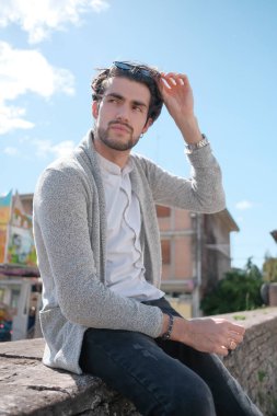 Yakışıklı, siyah saçlı, ceketli, güneşli bir günde alçak duvarda oturan adam. Yüksek kalite fotoğraf