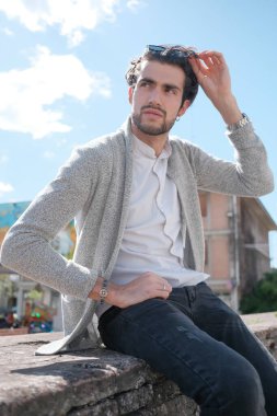 Yakışıklı, siyah saçlı, ceketli, güneşli bir günde alçak duvarda oturan adam. Yüksek kalite fotoğraf