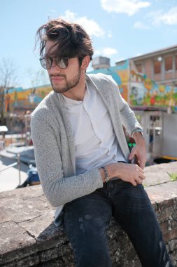 Yakışıklı, siyah saçlı, ceketli, güneşli bir günde alçak duvarda oturan adam. Yüksek kalite fotoğraf