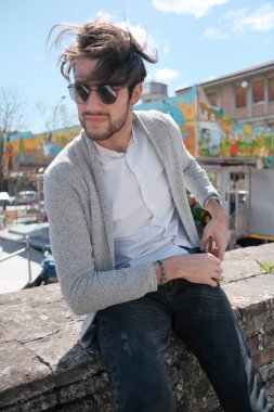 Yakışıklı, siyah saçlı, ceketli, güneşli bir günde alçak duvarda oturan adam. Yüksek kalite fotoğraf