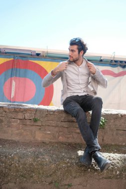 Yakışıklı, siyah saçlı, ceketli, güneşli bir günde alçak duvarda oturan adam. Yüksek kalite fotoğraf