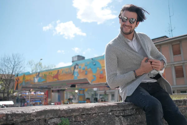 Yakışıklı, siyah saçlı, ceketli, güneşli bir günde alçak duvarda oturan adam. Yüksek kalite fotoğraf