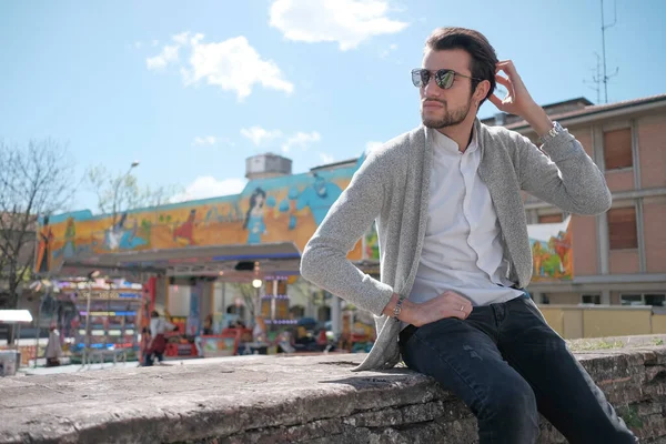 Yakışıklı, siyah saçlı, ceketli, güneşli bir günde alçak duvarda oturan adam. Yüksek kalite fotoğraf