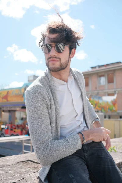 Yakışıklı, siyah saçlı, ceketli, güneşli bir günde alçak duvarda oturan adam. Yüksek kalite fotoğraf
