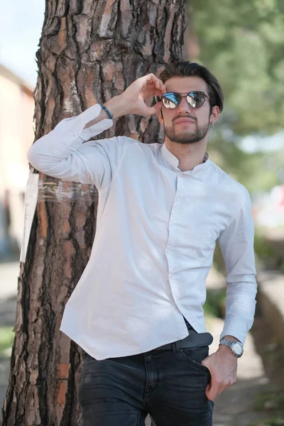 Yakışıklı, siyah saçlı, güneş gözlüklü, geniş çam gövdesine yaslanmış bir çocuk. Yüksek kalite fotoğraf