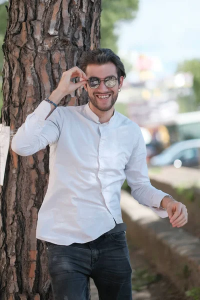 Yakışıklı, siyah saçlı, güneş gözlüklü, geniş çam gövdesine yaslanmış bir çocuk. Yüksek kalite fotoğraf