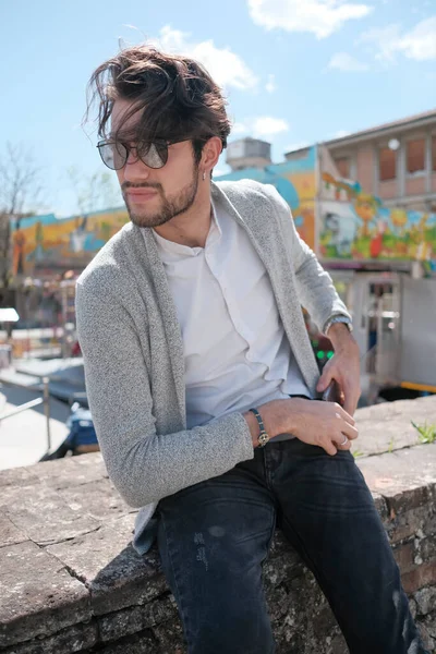 Yakışıklı, siyah saçlı, ceketli, güneşli bir günde alçak duvarda oturan adam. Yüksek kalite fotoğraf