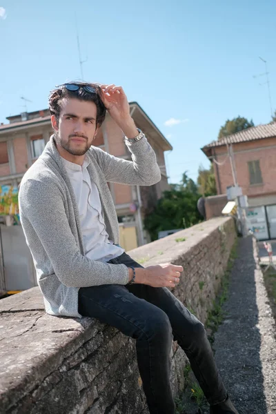 Yakışıklı, siyah saçlı, ceketli, güneşli bir günde alçak duvarda oturan adam. Yüksek kalite fotoğraf
