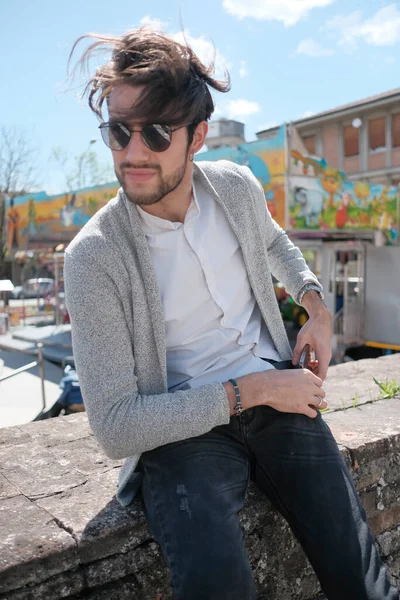Yakışıklı, siyah saçlı, ceketli, güneşli bir günde alçak duvarda oturan adam. Yüksek kalite fotoğraf