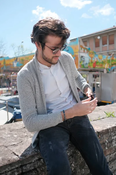 Yakışıklı, siyah saçlı, ceketli, güneşli bir günde alçak duvarda oturan adam. Yüksek kalite fotoğraf