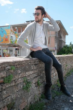 Yakışıklı, siyah saçlı, ceketli, güneşli bir günde alçak duvarda oturan adam. Yüksek kalite fotoğraf