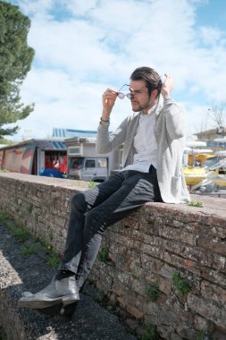 Yakışıklı, siyah saçlı, ceketli, güneşli bir günde alçak duvarda oturan adam. Yüksek kalite fotoğraf