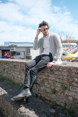 Yakışıklı, siyah saçlı, ceketli, güneşli bir günde alçak duvarda oturan adam. Yüksek kalite fotoğraf