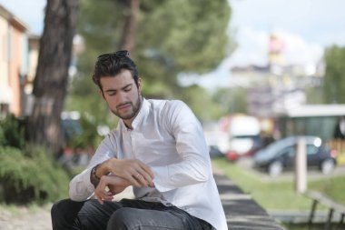 Güzel, siyah saçlı İtalyan çocuk ağaçların altında oturmuş saatini kontrol ediyor. Yüksek kalite fotoğraf