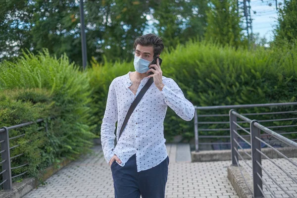 Yakışıklı, koyu renk saçlı İtalyan çocuk anti-covid maskeyle yürüyor ve telefonda konuşuyor. Yüksek kalite fotoğraf
