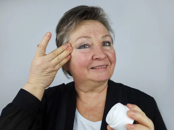 An elderly woman applies an anti-aging face cream. Cosmetology. Withering (aging) of the skin. Pigmentation. Couperose.