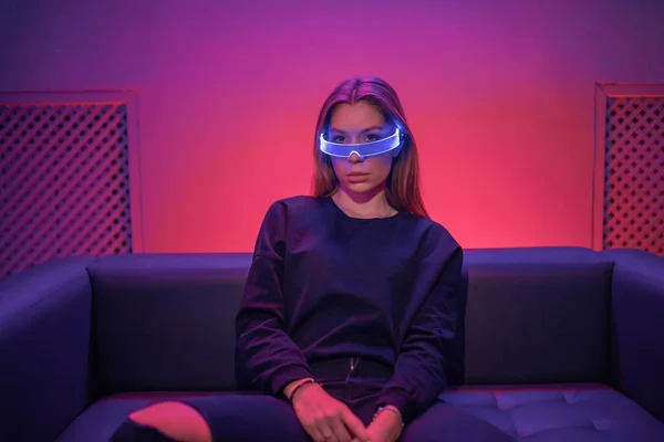 Neon noir. Mulher sentada em uma sala de néon usando óculos de néon. — Fotografia de Stock