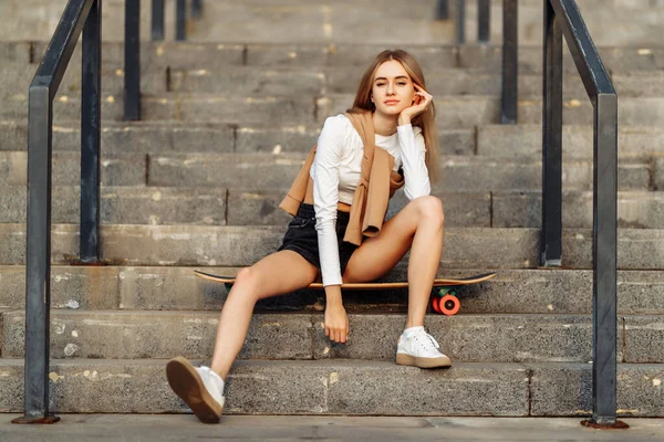 サングラスをかけて階段に腰掛けているロングボードを持つクールな若い女性。カジュアルスタイル — ストック写真