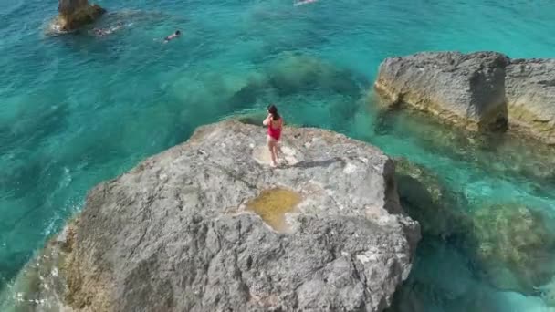 赤い水着姿の美人が海を眺めながら大きな岩を買う — ストック動画