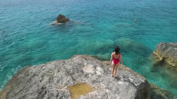 Beautiful woman in red swimsuit walking buy big rock looking at the sea — Stock Video