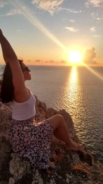 Mujer sentada en el acantilado disfrutando de la puesta de sol sobre el mar — Vídeos de Stock