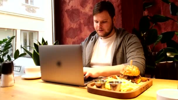 カフェでノートパソコンでバーガーを食べる男 — ストック動画