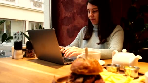Kvinne som jobber fjernstyrt på laptop i kafe-spisende burger drikke te – stockvideo